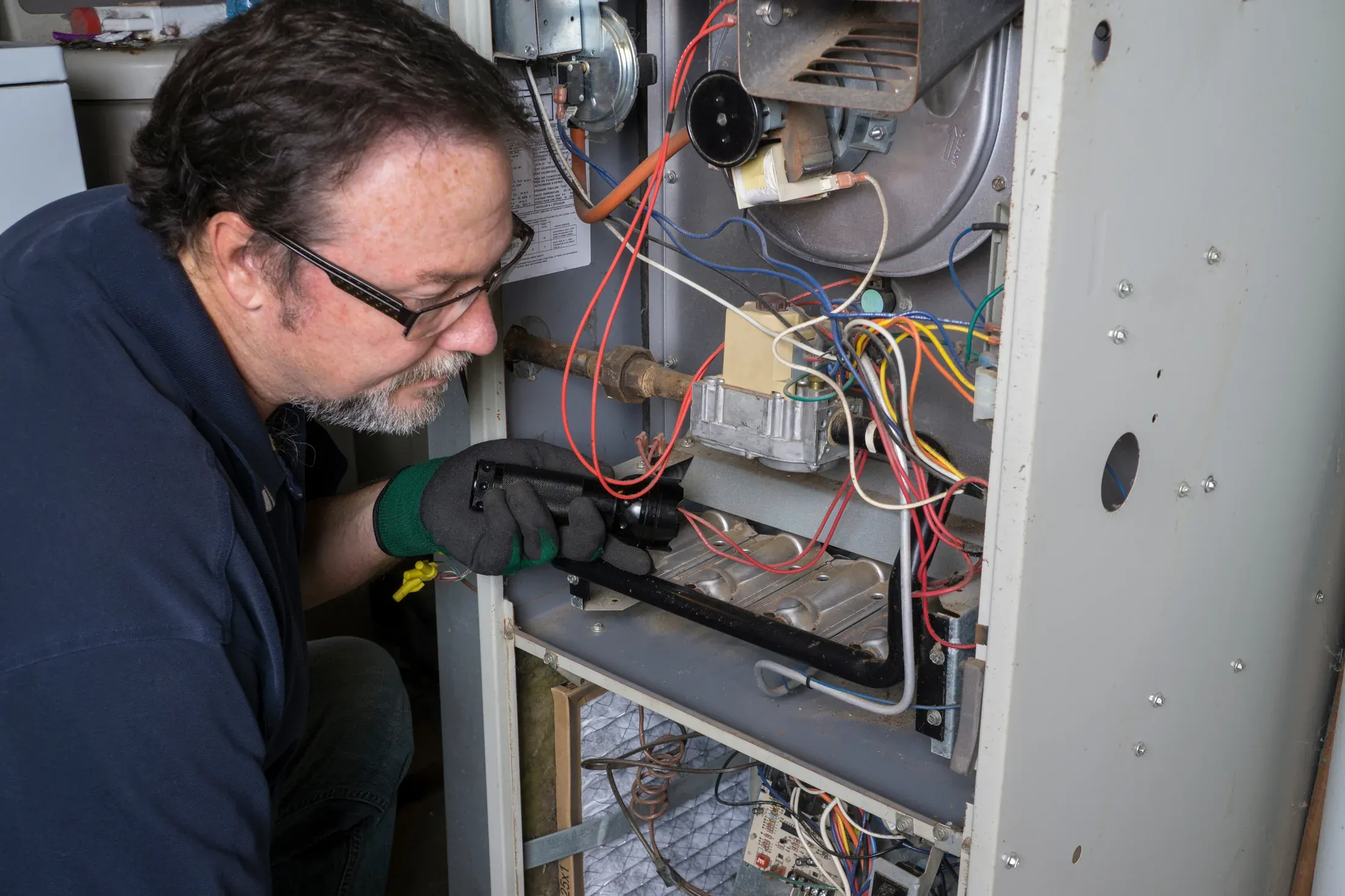 HVAC technician servicing an open residential gas furnace, inspecting the wiring and burner assembly
